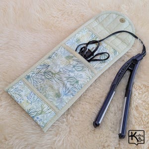 May include: A quilted fabric heat-resistant pouch featuring a light green and blue botanical print with a snap closure, containing a black hair straightener with its power cord coiled inside the open pocket, and a small logo in the corner.
