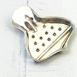 May include: A vintage, silver-toned citrus juicer. The juicer has a distinctive shape with a curved handle and a perforated pressing surface. Small, brass-coloured rivets are visible on the pressing surface. The juicer is designed for hand-squeezing citrus fruits.