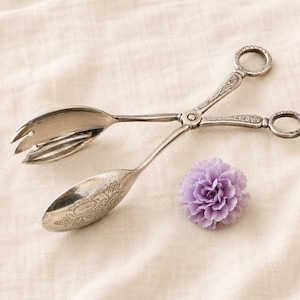 May include: A pair of silver-toned metal serving tongs with ring-shaped handles and intricate floral engravings on the arms. One side features a three-pronged fork end, while the other is a smooth spoon. A small, light purple artificial flower is nearby.