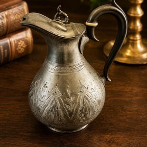 May include: A metallic pitcher with a tarnished silver finish, featuring an ornate engraved floral and geometric pattern on the body. It has a curved handle with a small light-coloured accent ring, a hinged lid with a finial, and a spout.