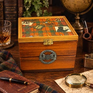 May include: A square wooden box with a hinged lid featuring a red and green tartan pattern decorated with illustrations of pipes and magnifying glasses. The front of the box displays a circular silver-toned metal emblem with a bow design.