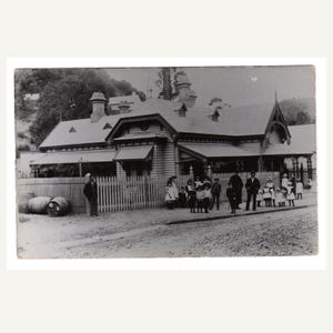 Op de afbeelding: Zwart-witfoto van een houten gebouw met een hek en een groep kinderen en volwassenen ervoor. Twee vaten zijn zichtbaar bij het hek. Het gebouw heeft een zadeldak en verschillende schoorstenen.