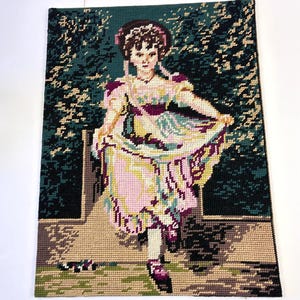 May include: Needlepoint tapestry depicting a young woman in a pink and yellow dress, holding her skirt. The background features a garden scene with green foliage and a stone wall. The artwork is framed with a dark green border.