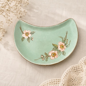May include: A crescent-shaped ceramic dish in a soft mint green hue, featuring hand-painted illustrations of delicate pink wild roses with green leaves and stems. The dish has a smooth, curved edge and a subtle speckled finish across its surface.