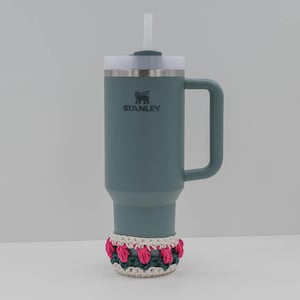 May include: A teal Stanley tumbler with a white lid and straw, featuring the Stanley logo. A handmade crochet cozy in white, pink, and green accents the base. The tumbler has a handle for easy carrying.