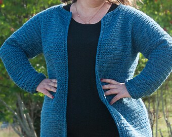 Op de afbeelding: Een tealblauwe gehaakte cardigan met lange mouwen en een open voorkant. De cardigan wordt gedragen over een zwart shirt en een zwarte broek.