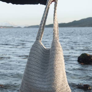 May include: A gray crocheted tote bag with long straps, held by a hand against a background of water and mountains.