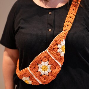 May include: Orange crochet crossbody bag with daisy flower details. The bag features a square pattern with white and yellow flower accents. The strap is also crocheted in orange. A stylish and unique handmade accessory.