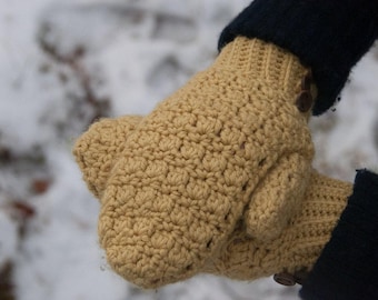 Peut inclure: Une paire de mitaines en crochet beige avec un motif de point texturé. Les mitaines ont un poignet côtelé et sont fixées avec un bouton marron. Les mitaines sont portées sur une manche bleu foncé, avec un fond enneigé.