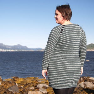 May include: A woman wearing a striped, blue and white sweater with black buttons. She is standing on a rocky shore with a body of water in the background.