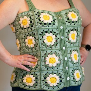 Peut inclure: Un haut en crochet sans manches composé de carrés granny vert sauge avec des motifs de marguerites blanches et jaunes. Le vêtement présente une encolure carrée, des bretelles larges et une patte de boutonnage avec six petits boutons blancs.