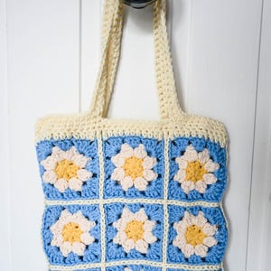 May include: A handmade crochet tote bag with a daisy pattern. The bag features a blue background with white and yellow flower details. The handles and top border are cream-colored. The bag is hanging on a black knob against a white background.