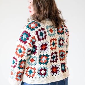 May include: A cream-colored, crocheted jacket featuring a patchwork design of colorful squares. The squares are in shades of teal, orange, burgundy, and cream. The jacket has long sleeves and a rounded neckline. The person is wearing blue jeans.