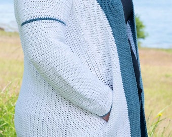 Op de afbeelding: Een wit en blauw gebreid vest met een lange mouw en zakken. Het vest is open aan de voorkant en heeft een blauwe streep aan de zijkant.