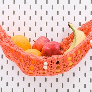 Puede incluir: Una hamaca para frutas tejida a ganchillo de color coral, suspendida por varillas de madera, con un patrón decorativo de tejido abierto y pequeñas cuentas de madera blanca. La hamaca contiene un plátano amarillo, dos manzanas rojas y dos naranjas.