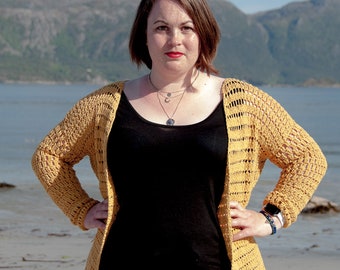 May include: A woman wearing a yellow crochet cardigan over a black tank top and blue jeans. She is standing on a sandy beach with mountains in the background.