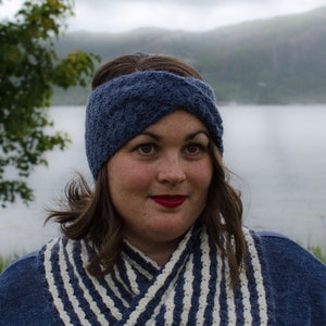 Puede incluir: Una mujer con una diadema de punto azul y una bufanda a rayas. La diadema es de color azul oscuro y parece estar hecha de un hilo suave. La bufanda tiene rayas blancas y azules. El fondo muestra un lago y montañas.