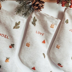 Pode incluir: Três meias de Natal bege com desenhos bordados. As meias estão penduradas em uma prateleira de madeira. As meias são decoradas com pequenas imagens bordadas de ramos de pinheiro, sinos, presentes e um chapéu de Papai Noel. As meias são personalizadas com os nomes "Joel", "Love" e "Joy".