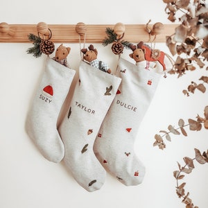 Pode incluir: Três meias de Natal brancas com desenhos bordados de ramos de pinheiro, bolotas e presentes. As meias estão penduradas em um suporte de madeira e cada uma tem um nome bordado na frente: Suzy, Taylor e Dulcie.