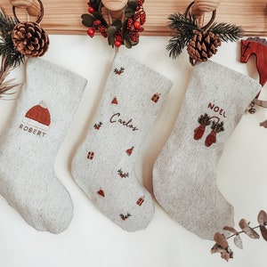 Pode incluir: Três meias de Natal brancas com os nomes bordados "Robert", "Carlos" e "Noel". Cada meia apresenta um pequeno desenho bordado, incluindo uma casa, uma rena e uma árvore de pinho.