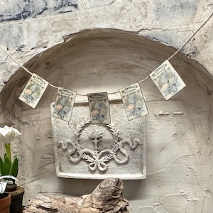 May include: A string of small, vintage-style cards with floral illustrations and text, strung across a textured, off-white wall. The cards are suspended above a decorative, carved stone planter with a botanical design.