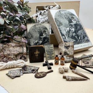 May include: A collection of miniature items including a wooden box with a black and white illustration, a small brown book with a gold cross, a wooden mallet, two glass vials, a wooden cross, two wooden stakes, and various printed paper documents.