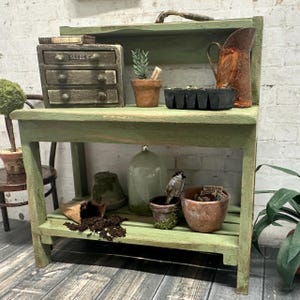 May include: A weathered, green potting bench with multiple shelves, holding miniature gardening supplies. Includes a small drawer unit labeled "SEEDS", terracotta pots, a rusty pitcher, and a glass cloche. The bench is set against a white brick wall.