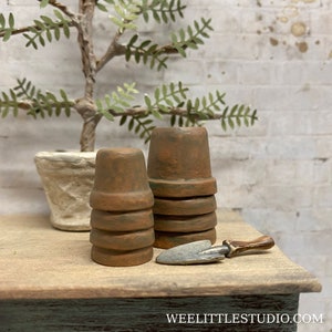 May include: Miniature terracotta flower pots stacked in two groups, with a small trowel. The pots are a warm brown color with subtle green accents. A small potted plant is in the background. The scene is set on a wooden surface.