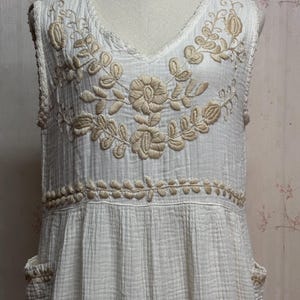 May include: A sleeveless white dress featuring a textured, crinkled fabric finish. The bodice is adorned with intricate beige floral embroidery around the V-neckline and a horizontal band at the waist. Two side pockets are visible at the hips.