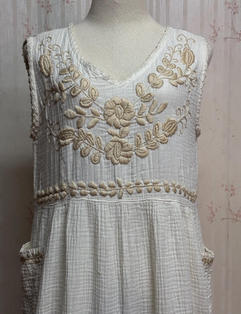 May include: A sleeveless white dress featuring a textured, crinkled fabric finish. The bodice is adorned with intricate beige floral embroidery around the V-neckline and a horizontal band at the waist. Two side pockets are visible at the hips.