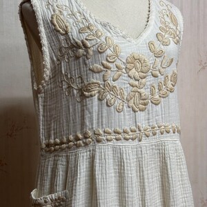 May include: A sleeveless off-white dress made of textured crinkled fabric featuring a V-neckline. The bodice is adorned with intricate beige floral embroidery around the neckline and a horizontal band at the waist, including a small side pocket.