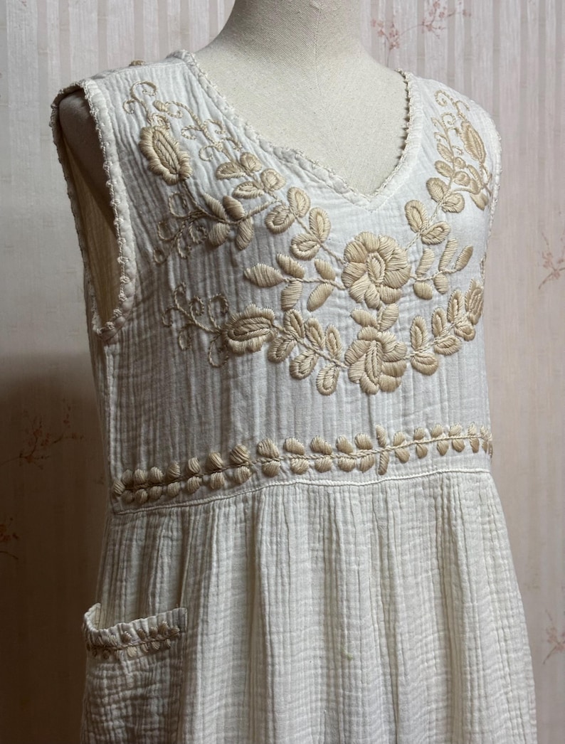 May include: A sleeveless off-white dress made of textured crinkled fabric featuring a V-neckline. The bodice is adorned with intricate beige floral embroidery around the neckline and a horizontal band at the waist, including a small side pocket.