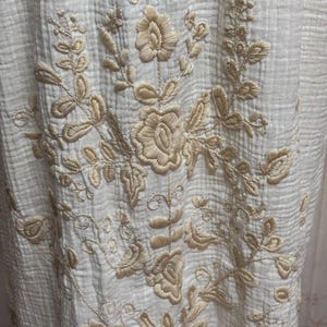 May include: A cream-colored fabric with a crinkled texture featuring intricate beige floral embroidery. The design consists of central rose-like blossoms surrounded by detailed leaves and scrolling vines, creating a symmetrical botanical pattern.