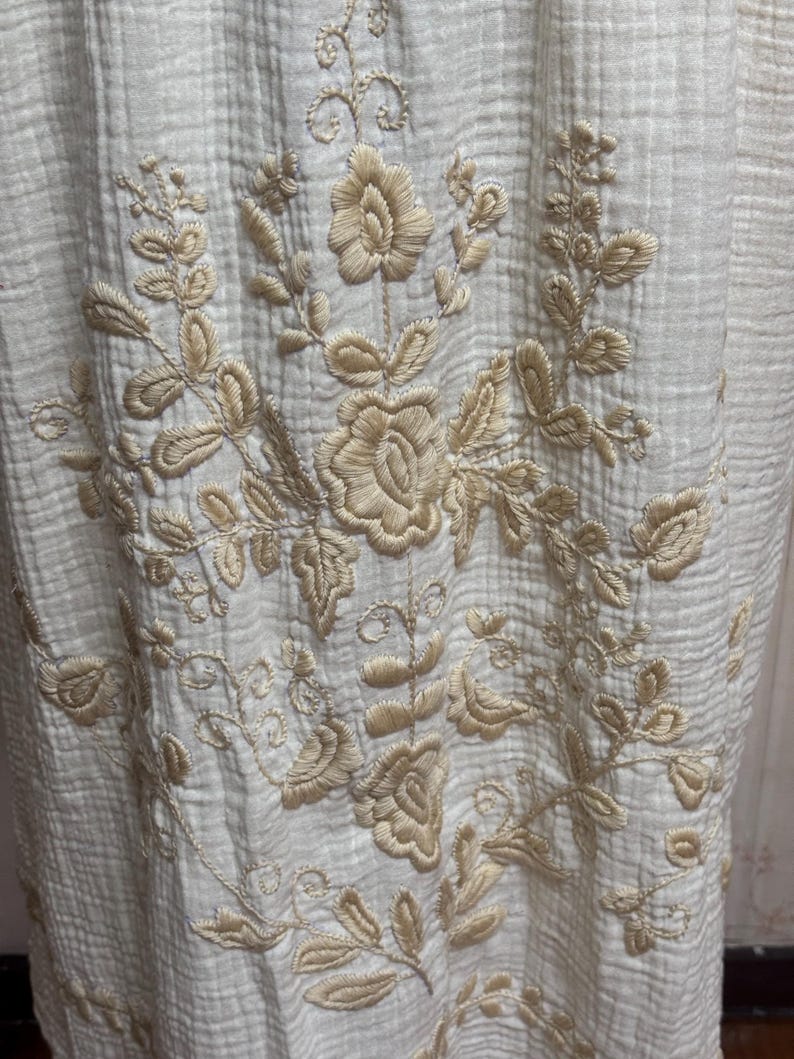 May include: A cream-colored fabric with a crinkled texture featuring intricate beige floral embroidery. The design consists of central rose-like blossoms surrounded by detailed leaves and scrolling vines, creating a symmetrical botanical pattern.