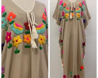 May include: A beige kaftan dress featuring vibrant, multi-colored floral embroidery across the chest and scattered throughout the garment. The dress includes a V-neckline with a white braided drawstring and tassels, plus white pom-pom trim along the sides.