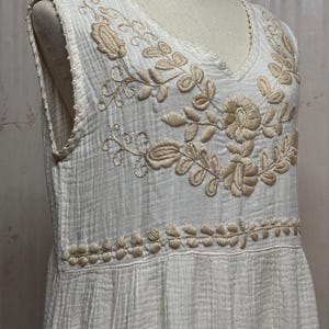 May include: A sleeveless white dress featuring a textured, crinkled fabric. The bodice is adorned with beige floral embroidery around the V-neckline and a horizontal band of stitched leaf patterns. A small patch pocket is visible on the lower side.