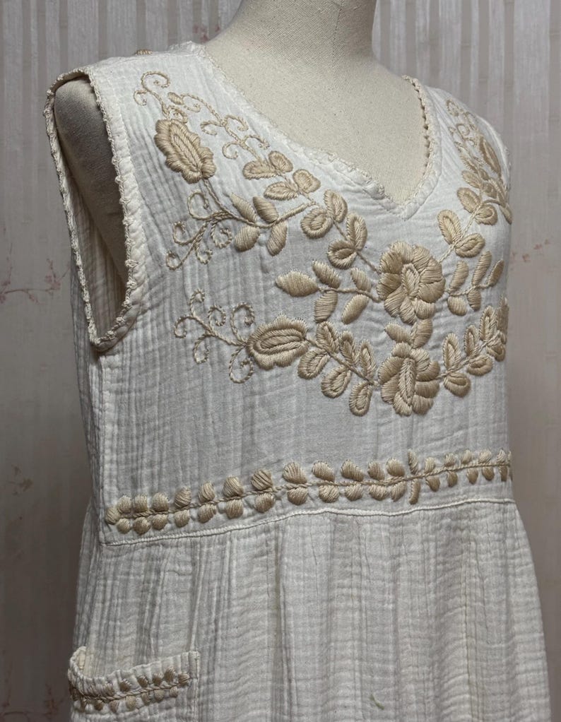 May include: A sleeveless white dress featuring a textured, crinkled fabric. The bodice is adorned with beige floral embroidery around the V-neckline and a horizontal band of stitched leaf patterns. A small patch pocket is visible on the lower side.