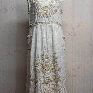 May include: A sleeveless white midi dress featuring a textured crinkle fabric and beige floral embroidery on the bodice and skirt. The dress includes a V-neckline, a defined waistline with horizontal embroidery, and two side pockets on the skirt.