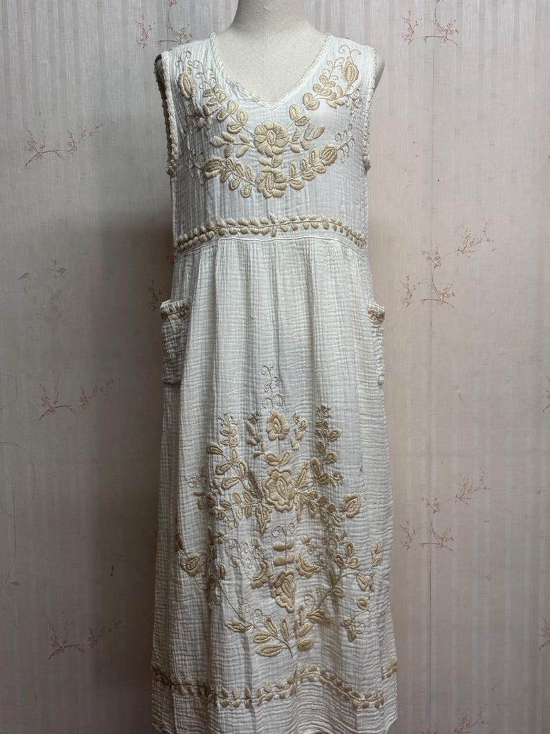May include: A sleeveless white midi dress featuring a textured crinkle fabric and beige floral embroidery on the bodice and skirt. The dress includes a V-neckline, a defined waistline with horizontal embroidery, and two side pockets on the skirt.