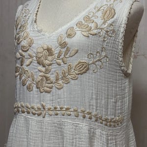 May include: A sleeveless white dress featuring a textured, crinkled fabric. The bodice is adorned with beige floral embroidery around the V-neckline and a horizontal band of leaf-patterned embroidery across the waistline area.