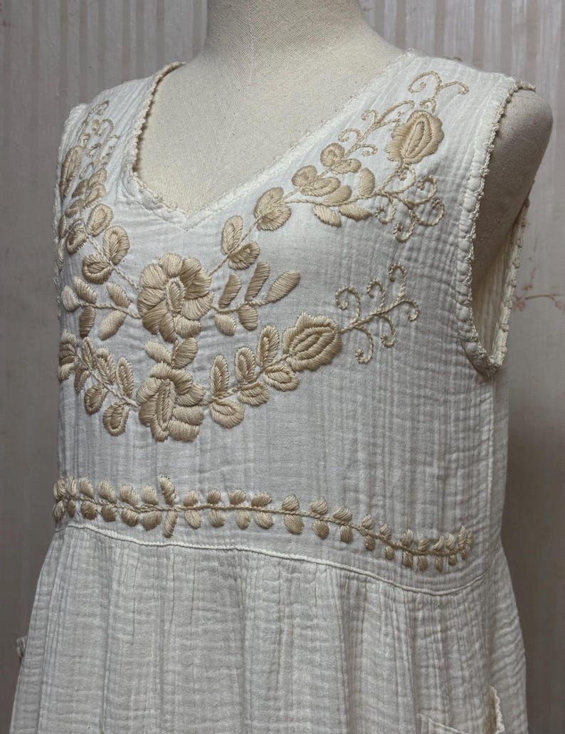 May include: A sleeveless white dress featuring a textured, crinkled fabric. The bodice is adorned with beige floral embroidery around the V-neckline and a horizontal band of leaf-patterned embroidery across the waistline area.
