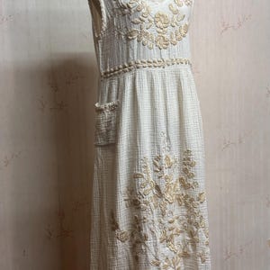 May include: A sleeveless, off-white midi dress featuring a textured, crinkled fabric. The dress is adorned with beige floral embroidery around the V-neckline, waist, and lower skirt, and includes a visible patch pocket on the right side.