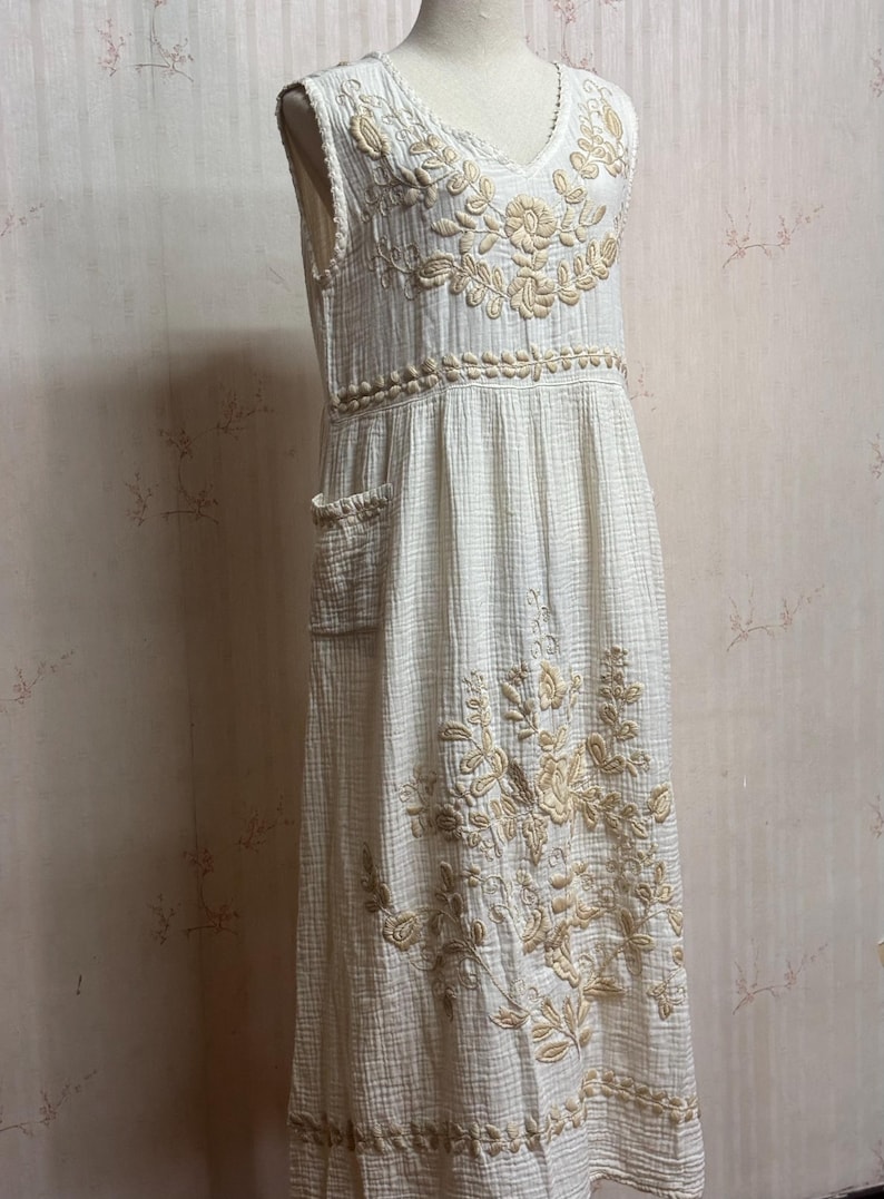 May include: A sleeveless, off-white midi dress featuring a textured, crinkled fabric. The dress is adorned with beige floral embroidery around the V-neckline, waist, and lower skirt, and includes a visible patch pocket on the right side.