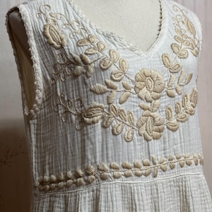 May include: A sleeveless white dress featuring a textured, crinkled fabric and a V-neckline. The bodice is adorned with intricate beige floral embroidery around the neckline and a horizontal band of leaf-like stitching across the waist area.
