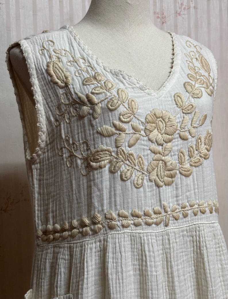May include: A sleeveless white dress featuring a textured, crinkled fabric and a V-neckline. The bodice is adorned with intricate beige floral embroidery around the neckline and a horizontal band of leaf-like stitching across the waist area.