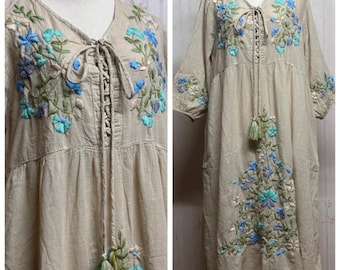 May include: A beige midi dress featuring a V-neckline with a lace-up front closure and long tassels. The dress is decorated with floral embroidery in shades of blue, turquoise, and green across the chest, sleeves, and lower front section.