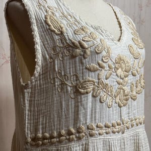 May include: A sleeveless white dress made of textured crinkled fabric featuring a V-neckline. The bodice is decorated with beige floral embroidery around the neckline and a horizontal band of leaf-like stitching across the waist area.