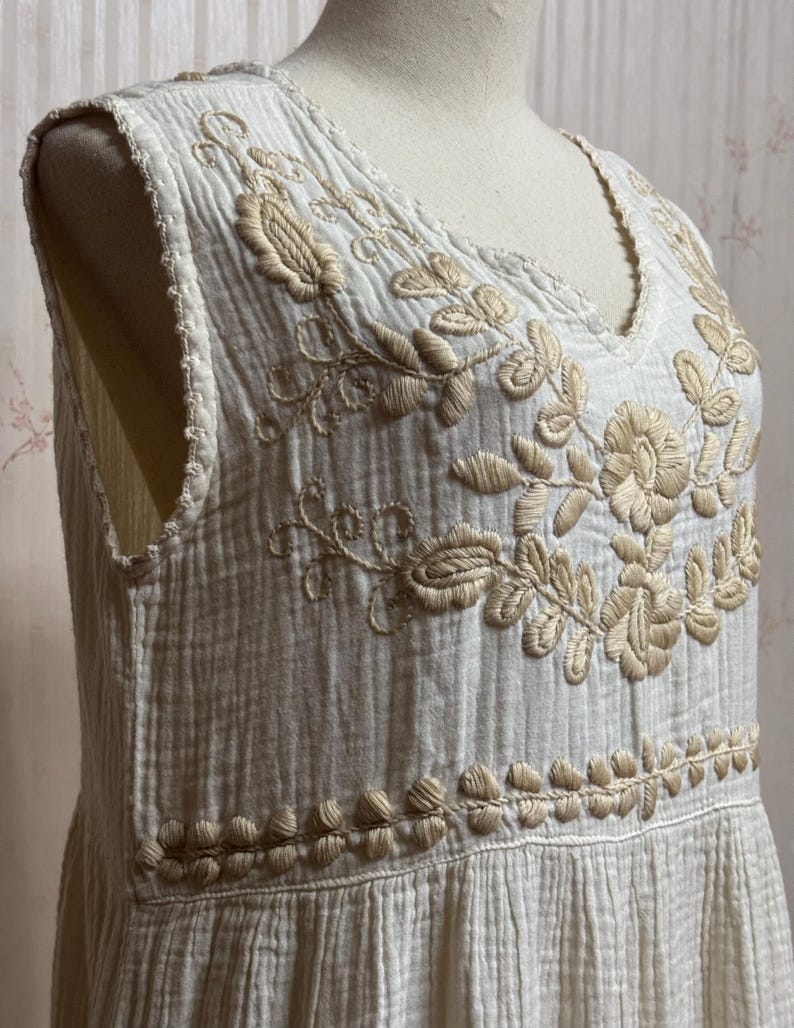May include: A sleeveless white dress made of textured crinkled fabric featuring a V-neckline. The bodice is decorated with beige floral embroidery around the neckline and a horizontal band of leaf-like stitching across the waist area.