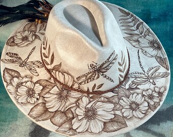 Pode incluir: Um chapéu fedora creme com aba larga, adornado com desenhos florais e de libélulas pintados à mão em tons de castanho. O chapéu apresenta uma faixa trançada castanha e penas decorativas.