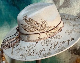 Pode incluir: Um chapéu fedora creme com um grande desenho de libélula. O chapéu apresenta uma faixa de couro castanho, uma borda trançada e um pequeno pingente de coração. A aba é decorada com ilustrações florais e de libélulas, e o texto "Ezer Kenegdo".